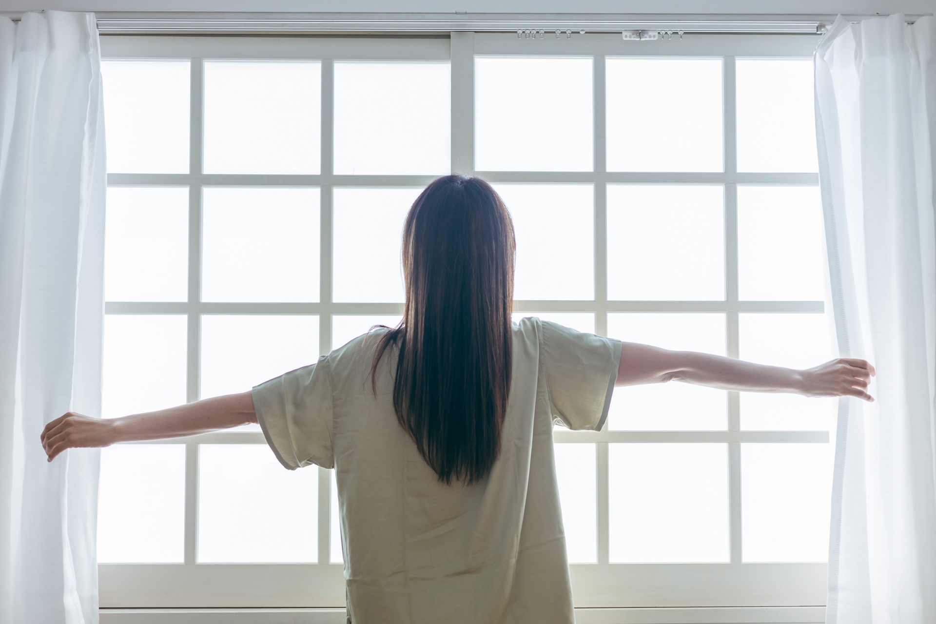 女性がカーテンを開けて朝日を浴びている後ろ姿